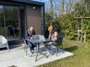 Werken vanaf de camping. Twee collega's aan een tuintafeltje met dikke jas, das en zonnebril op in een digitale meeting.